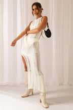 Woman in a white sleeveless maxi dress with a high slit and sheer knit, holding a black and white handbag, with cream boots.