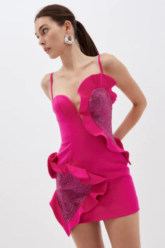Model wearing a vibrant pink mini dress with asymmetrical ruffles and rhinestone embellishments, posing on a white background.