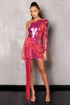 Woman wearing a pink and purple sequined one-shoulder mini dress with an asymmetrical hemline and a long draping detail, standing against a grey textured wall, full body view.