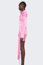 Side view of a model wearing a pink long-sleeve mini dress with asymmetrical draping and a twist detail at the neckline. The dress has a cutout detail at the chest and a ring hardware detail on the hip. The hem is asymmetrical with a draped panel.