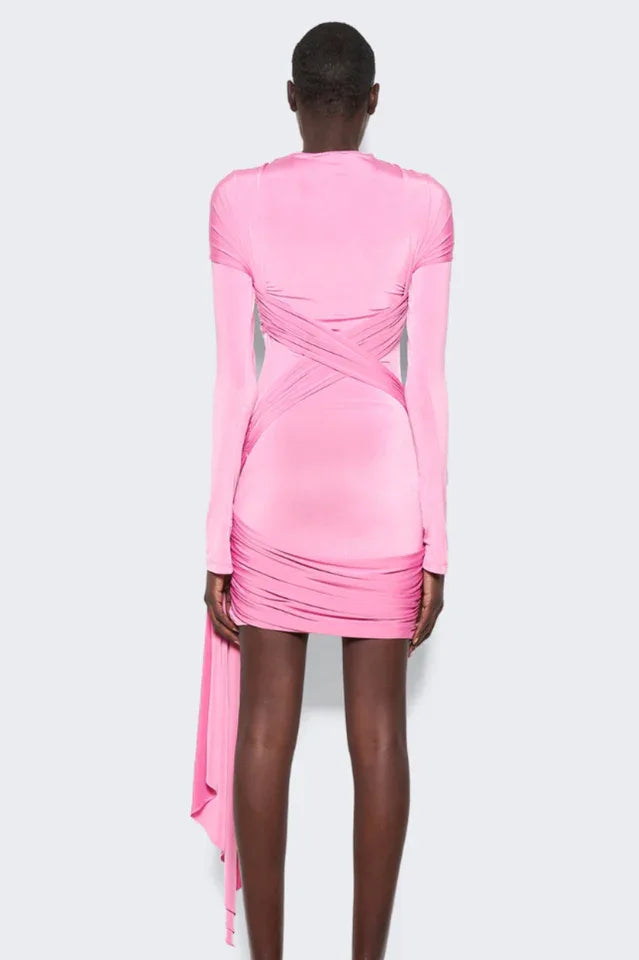 Back view of a model wearing a pink long-sleeve mini dress with asymmetrical draping and a twist detail at the neckline. The dress has a cutout detail at the chest and a ring hardware detail on the hip. The hem is asymmetrical with a draped panel.