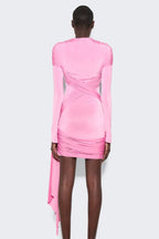 Back view of a model wearing a pink long-sleeve mini dress with asymmetrical draping and a twist detail at the neckline. The dress has a cutout detail at the chest and a ring hardware detail on the hip. The hem is asymmetrical with a draped panel.