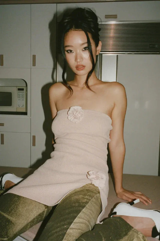 Model wearing a light pink strapless mini dress with floral embellishments and gold textured pants, seated on a kitchen counter.