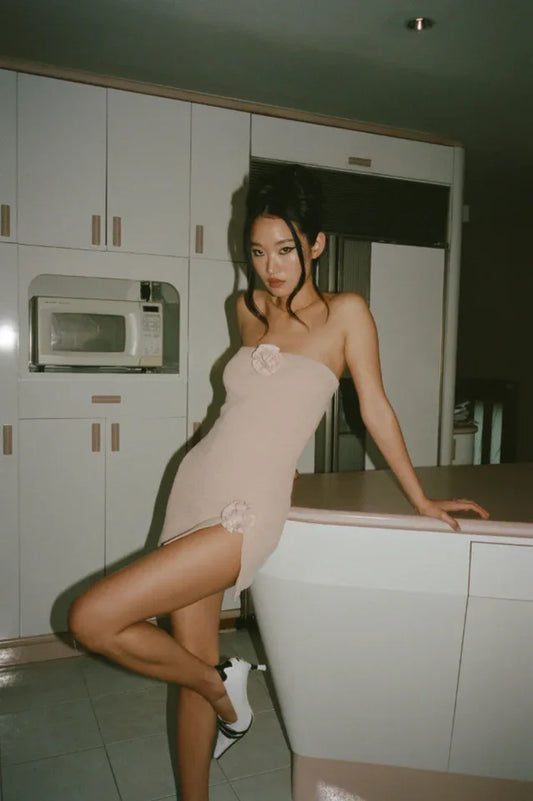 Model in a light pink strapless mini dress with floral embellishments and gold textured pants, leaning on a kitchen counter.