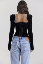 Back view of a model wearing a black velvet long-sleeve top with corset-style lacing, paired with light-wash denim jeans.