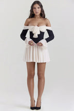 Woman in a black and ivory off-the-shoulder mini dress with a bow and flared sleeves, standing with hands clasped.