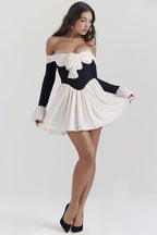 Woman in a black and ivory off-the-shoulder mini dress with a bow and flared sleeves, posing with one leg forward.