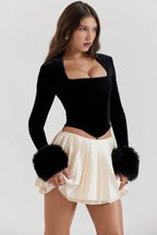 Close-up of a woman's torso wearing a black velvet top with faux fur cuffs and a cream pleated mini skirt.