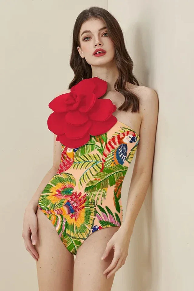 Woman wearing a one-piece swimsuit with a tropical parrot and palm leaf print, featuring a large red floral applique on the left shoulder, leaning against a wall. Main image