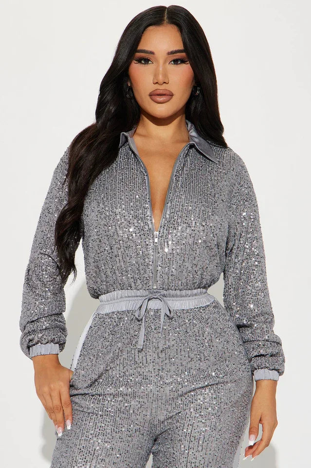 Close-up front view of a woman wearing a grey sequined long-sleeved collared jumpsuit with a zipper front closure and drawstring waist.