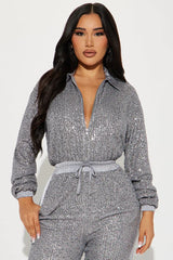 Close-up front view of a woman wearing a grey sequined long-sleeved collared jumpsuit with a zipper front closure and drawstring waist.