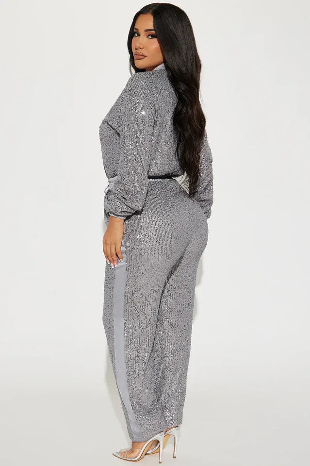 Side view of a woman wearing a grey sequined long-sleeved collared jumpsuit with a zipper front closure and drawstring waist. The pants have a grey side stripe.