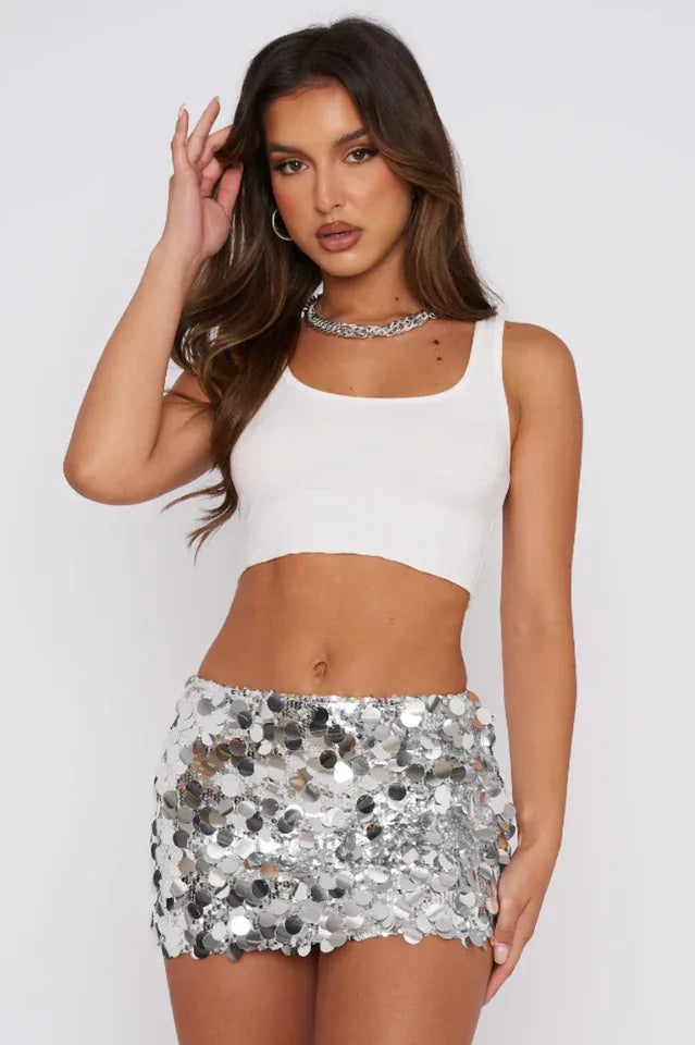 Woman wearing a white crop top and a silver sequined mini skirt, posing with her hand on her head. Main image