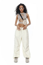 Full-body shot of a model wearing a taupe sleeveless cropped top with cut-outs, light-colored baggy cargo pants, and sneakers.