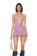 Woman in a lavender denim halter mini dress with a plunging neckline, pleated skirt, and denim boots, holding a silver handbag.