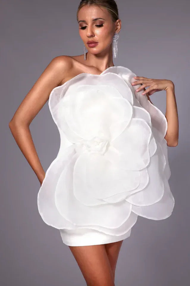 Woman wearing a white one-shoulder mini dress with a large floral appliqué on the bodice, close-up.