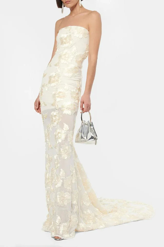 Woman wearing a strapless ivory gown with gold floral embroidery and a train, holding a silver handbag.