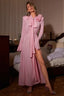Woman wearing a pink maxi dress with a bow neck, flared sleeves, and a high slit, standing in a bedroom.