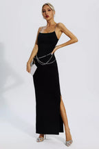 Woman wearing a black sleeveless maxi dress with chain straps and a chain belt, holding a clutch, posing.
