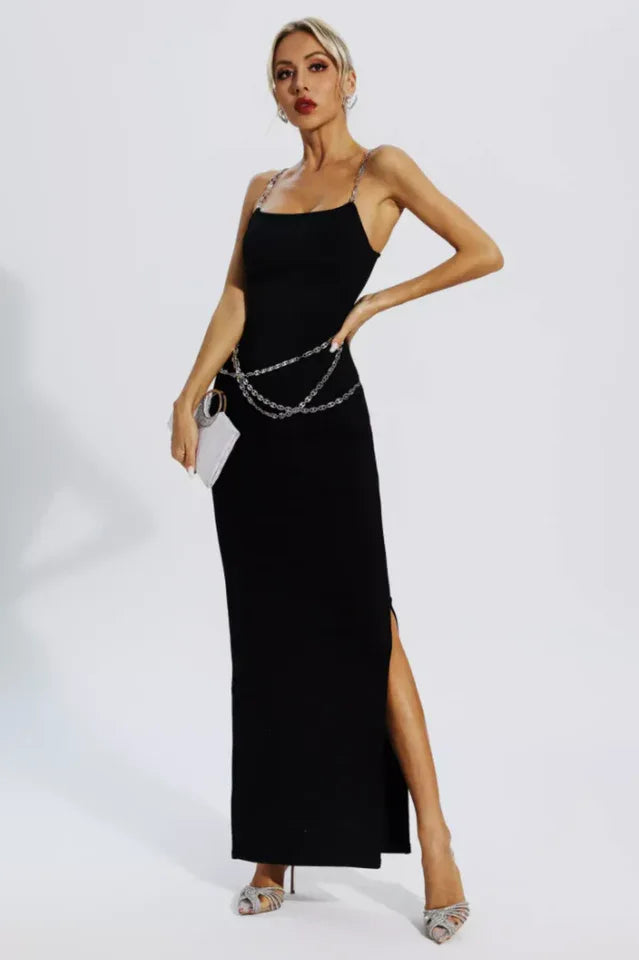 Woman wearing a black sleeveless maxi dress with chain straps and a chain belt, holding a clutch, posing.