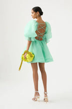 Back view of a model wearing a light green organza mini dress with voluminous puff sleeves, a ruched bodice, and an open back with criss-cross lacing, holding a lime green clutch.