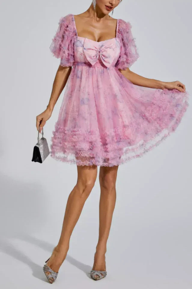 Woman in a pink floral print tulle dress with puff sleeves and bow detail, holding a silver clutch.