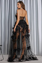 A woman poses in a dramatic black formal gown featuring a strapless bodice and a voluminous, sheer skirt adorned with flowers