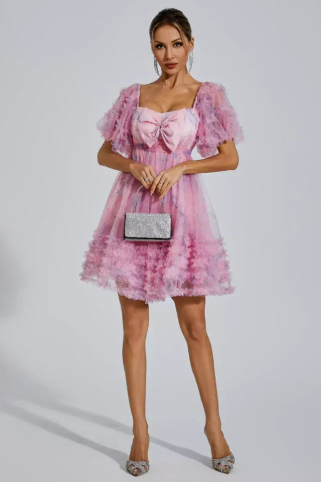 Woman in a pink floral print tulle dress with puff sleeves, bow detail, holding a silver clutch and wearing heels.
