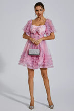 Woman in a pink floral print tulle dress with puff sleeves, bow detail, holding a silver clutch and wearing heels.