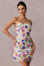 Model wearing a white mini slip dress adorned with vibrant floral sequins and gold chain straps, with a neutral background.