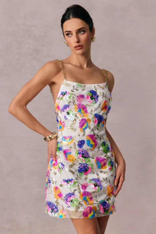 Model wearing a white mini slip dress adorned with vibrant floral sequins and gold chain straps, with a neutral background.