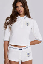 Front view of a model wearing a white cropped polo shirt with blue trim and matching white shorts with blue trim, displaying embroidered logos.