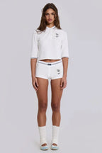 Full body shot of a model wearing a white cropped polo shirt, white shorts, and white socks with light blue ballet flats.