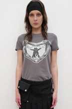 Model wearing a grey crew neck t-shirt with a black and white graphic print on the chest, a black belt bag, and a black pleated skirt.