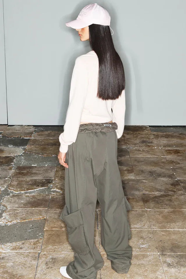 Rear view of a model wearing a distressed cream-colored cropped sweater and wide-leg olive green cargo pants, with a pink baseball cap.