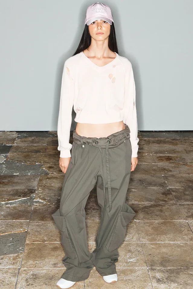 Model wearing a distressed cream-colored cropped sweater and wide-leg olive green cargo pants, with a pink baseball cap.
