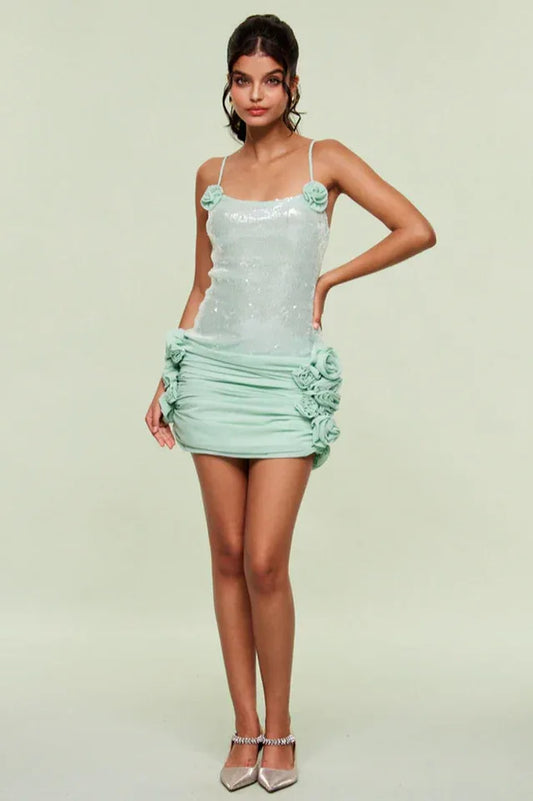 Full body shot of a woman in a light blue sequined mini dress with floral appliques and ruched detailing, standing with hands on hips.