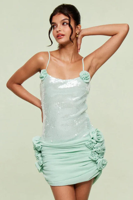 Woman wearing a light blue sequined mini dress with floral appliques and ruched detailing, posed with hand on hip.