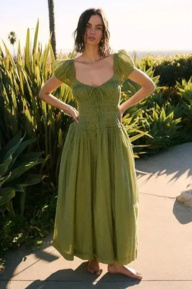 Woman wearing a green puff-sleeve midi dress with a smocked bodice and flowing skirt, standing outdoors with lush greenery in the background.