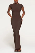 Woman wearing a dark brown full-length ribbed knit dress with short sleeves and a crew neck, standing with hands on hips against a white background.