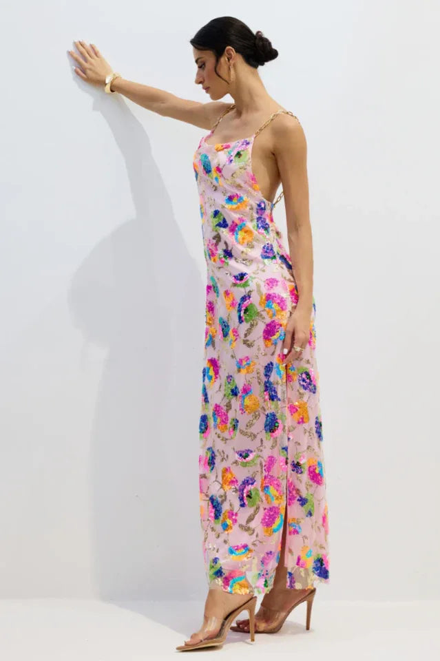 Side view of a woman in a pink sequined floral gown with gold chain straps, leaning against a white wall.