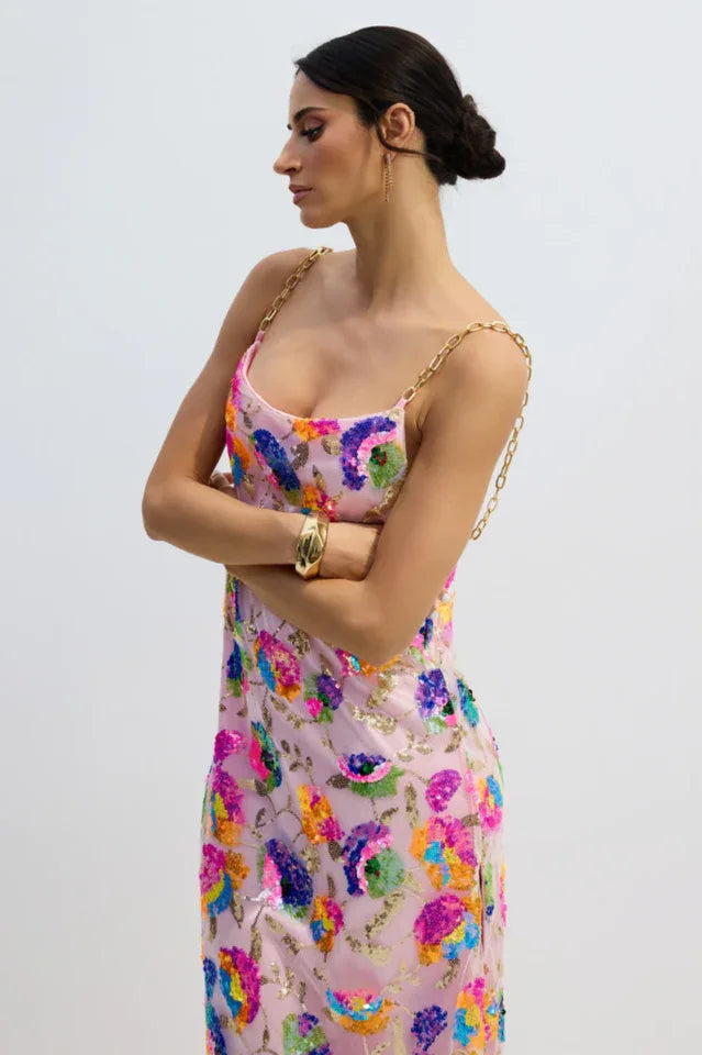 Woman in a pink sequined floral gown with gold chain straps, arms crossed.
