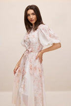 Woman posing in a floral print dress with sheer overlay.