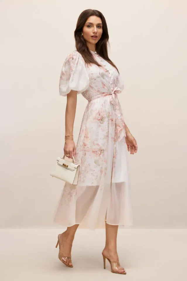Woman walking in a floral dress with a white handbag and clear heels.