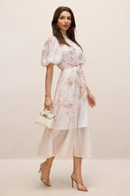 Woman walking in a floral dress with a white handbag and clear heels.