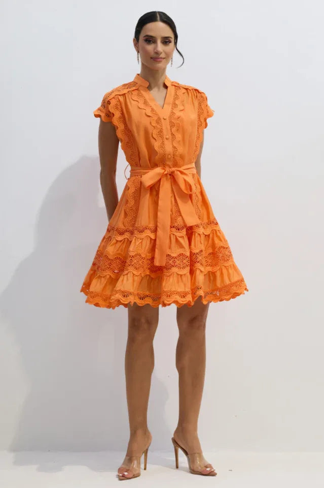 Woman standing and modeling an orange lace dress with a tie waist and clear heels.