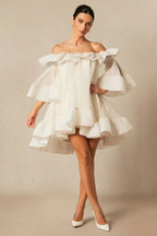 Model wearing a white off-the-shoulder dress with voluminous ruffles and bell sleeves, standing with legs crossed and hands clasped.