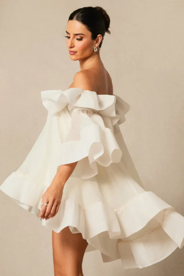 Model wearing a white off-the-shoulder dress with voluminous ruffles and bell sleeves, turning to the side.