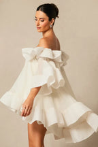 Model wearing a white off-the-shoulder dress with voluminous ruffles and bell sleeves, turning to the side.