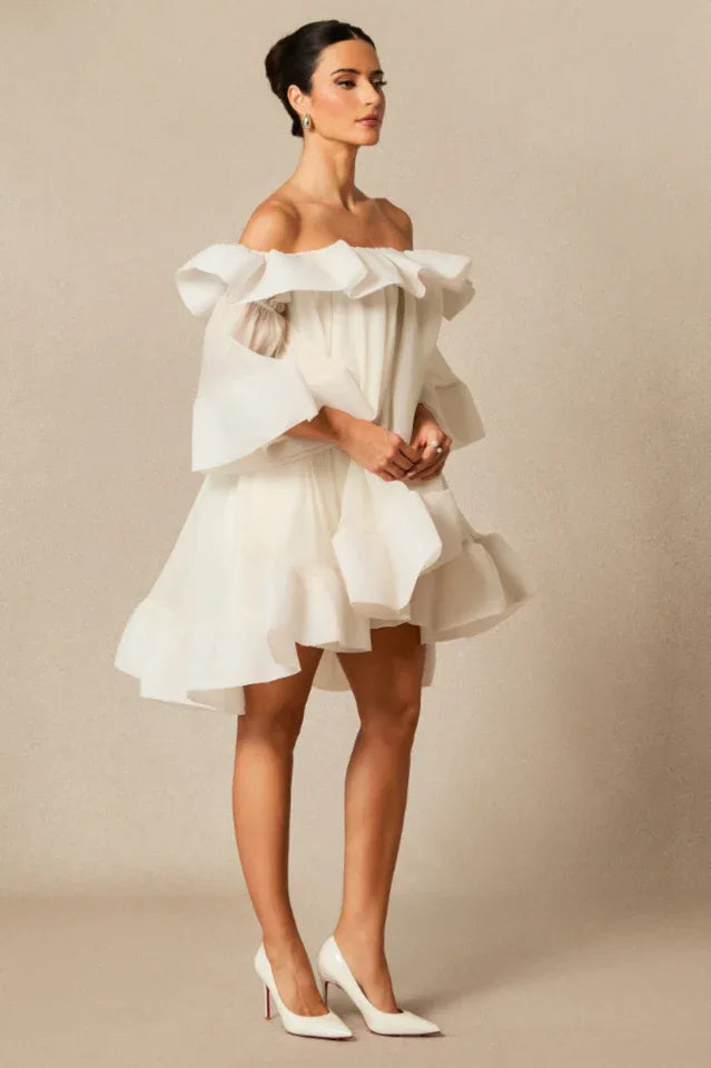 Model wearing a white off-the-shoulder dress with voluminous ruffles and bell sleeves, standing in profile with hands clasped.